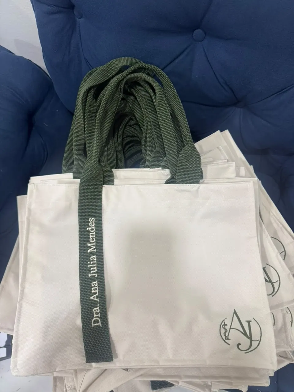 Pilha de bolsas cru com alças verde militar bordadas “Dra. Ana Julia Mendes”
