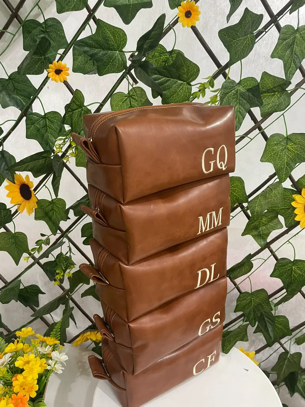 Cinco necessaires caramelo empilhadas, cada uma com iniciais diferentes bordadas